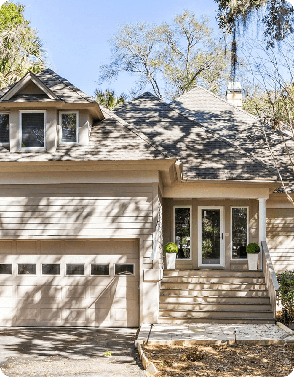 Two-story house with a beige exterior, a garage, and a central entrance with double doors. The front steps are flanked by potted plants. Trees and sunlight cast shadows on the scene.