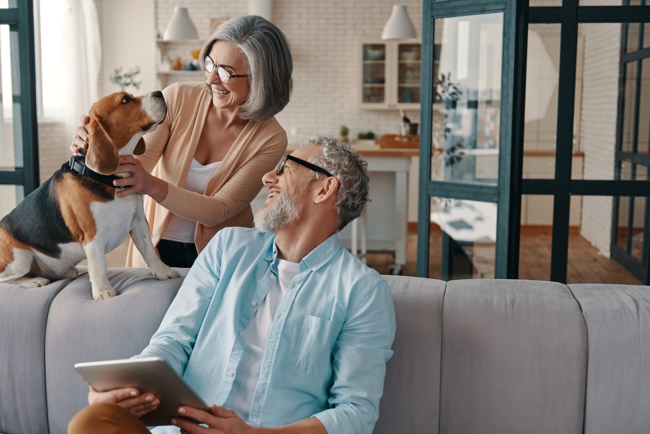 An older couple sits on a couch with a beagle. The woman pets the dog while the man, holding a tablet, looks at them, both smiling.