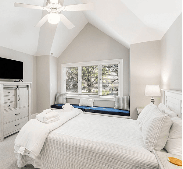 Bright bedroom with a sloped ceiling, ceiling fan, and large window. Features a bed, dresser with a TV, and a window seat with cushions. Neutral-colored decor and neatly arranged linens.