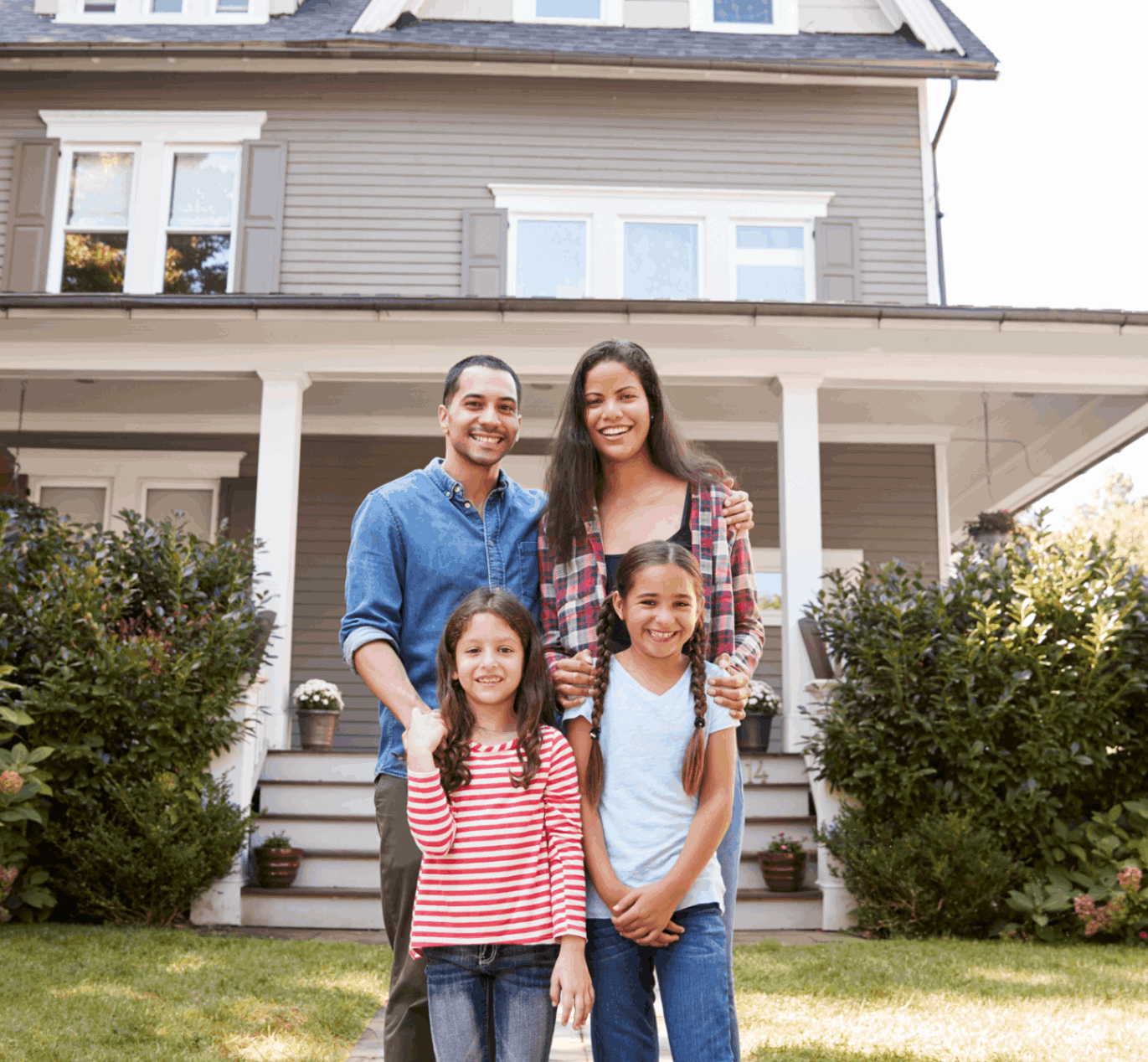 A family of four stands smiling in front of a two-story house. The parents are behind the two children, with greenery and a porch visible in the background.