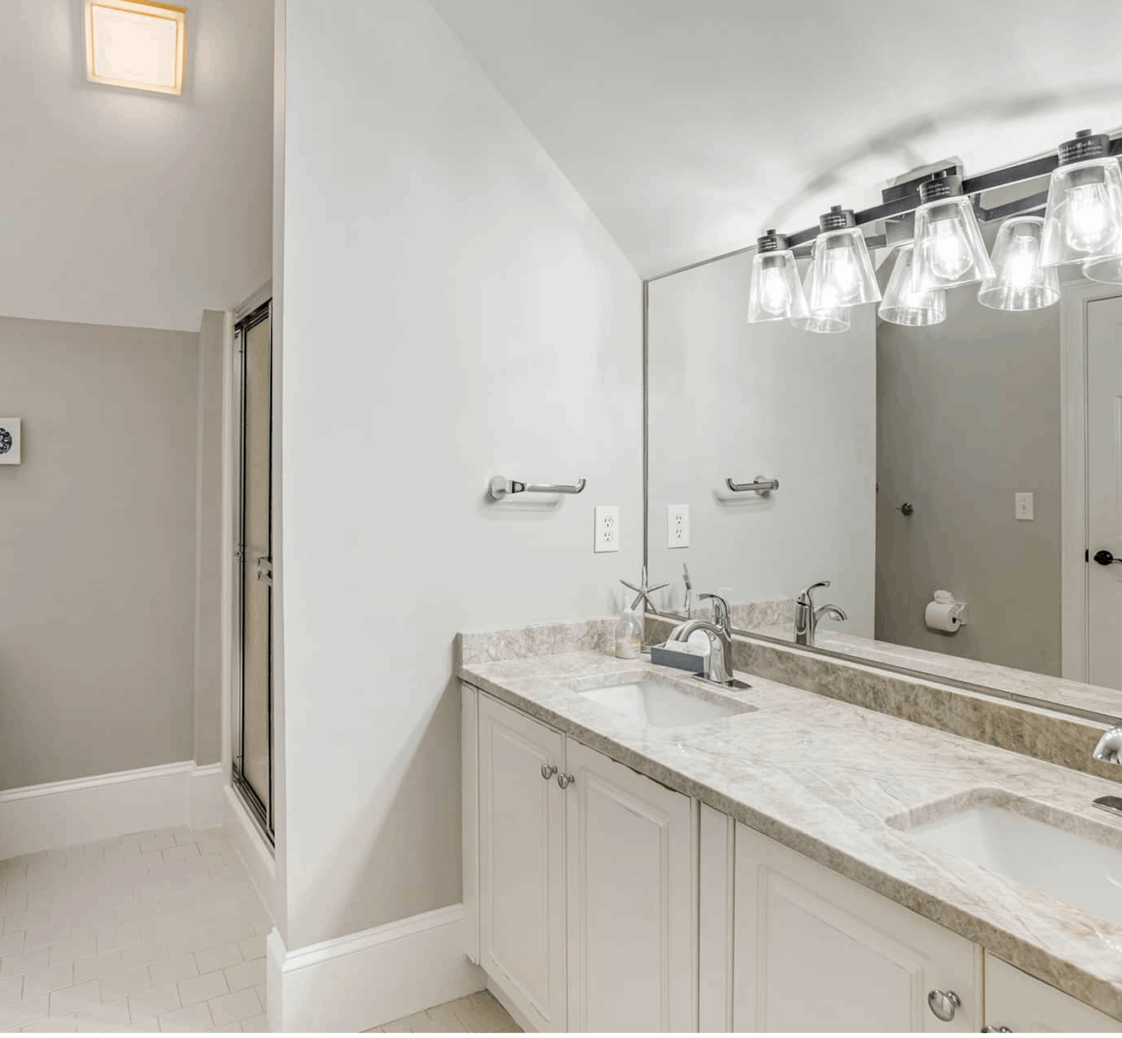 Bathroom with a marble countertop, dual sinks, large mirror, and modern light fixtures. There's a glass shower door and light gray walls.