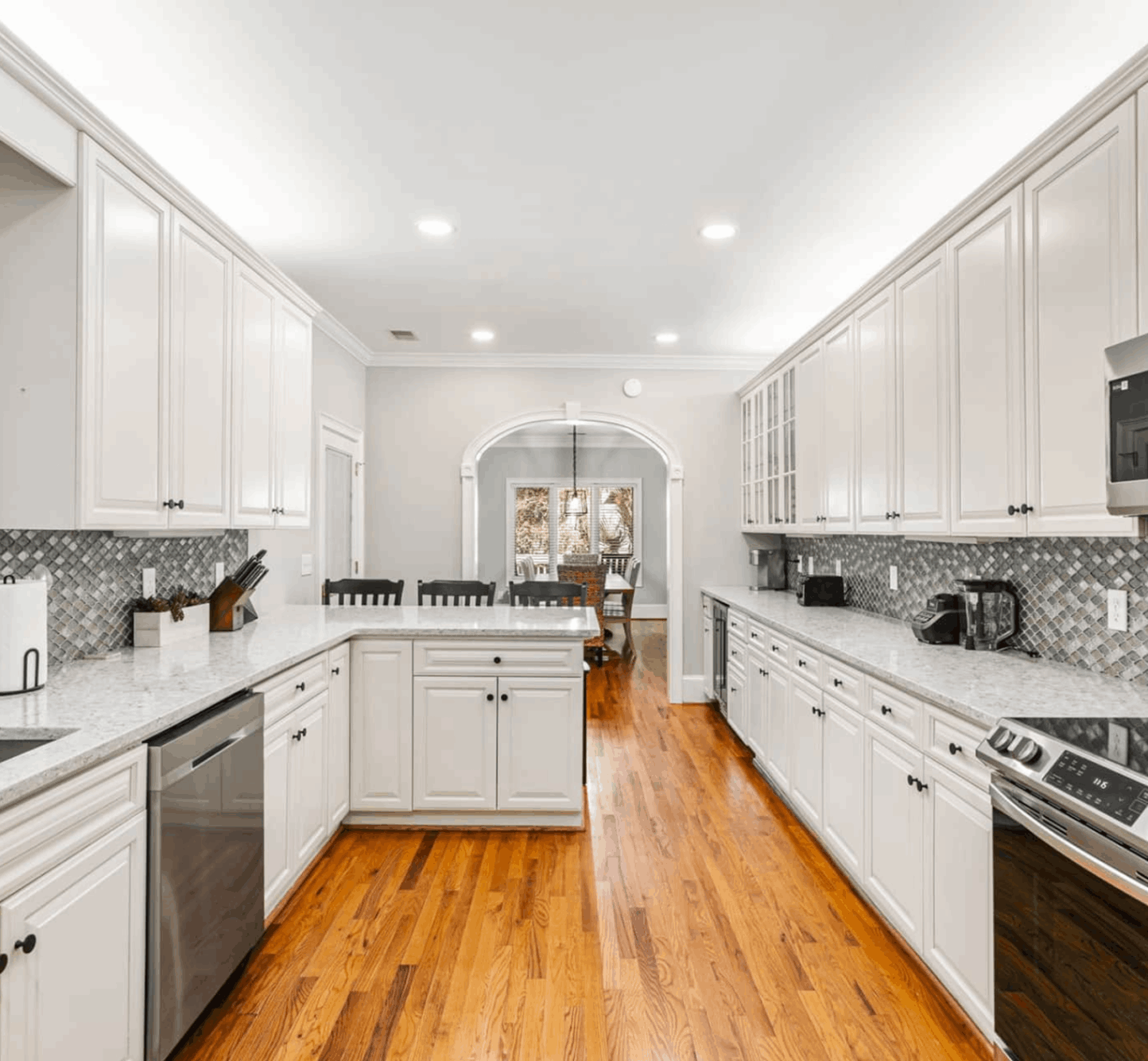 A spacious kitchen with white cabinets, wood flooring, stainless steel appliances, and a tile backsplash. An archway leads to a dining room.