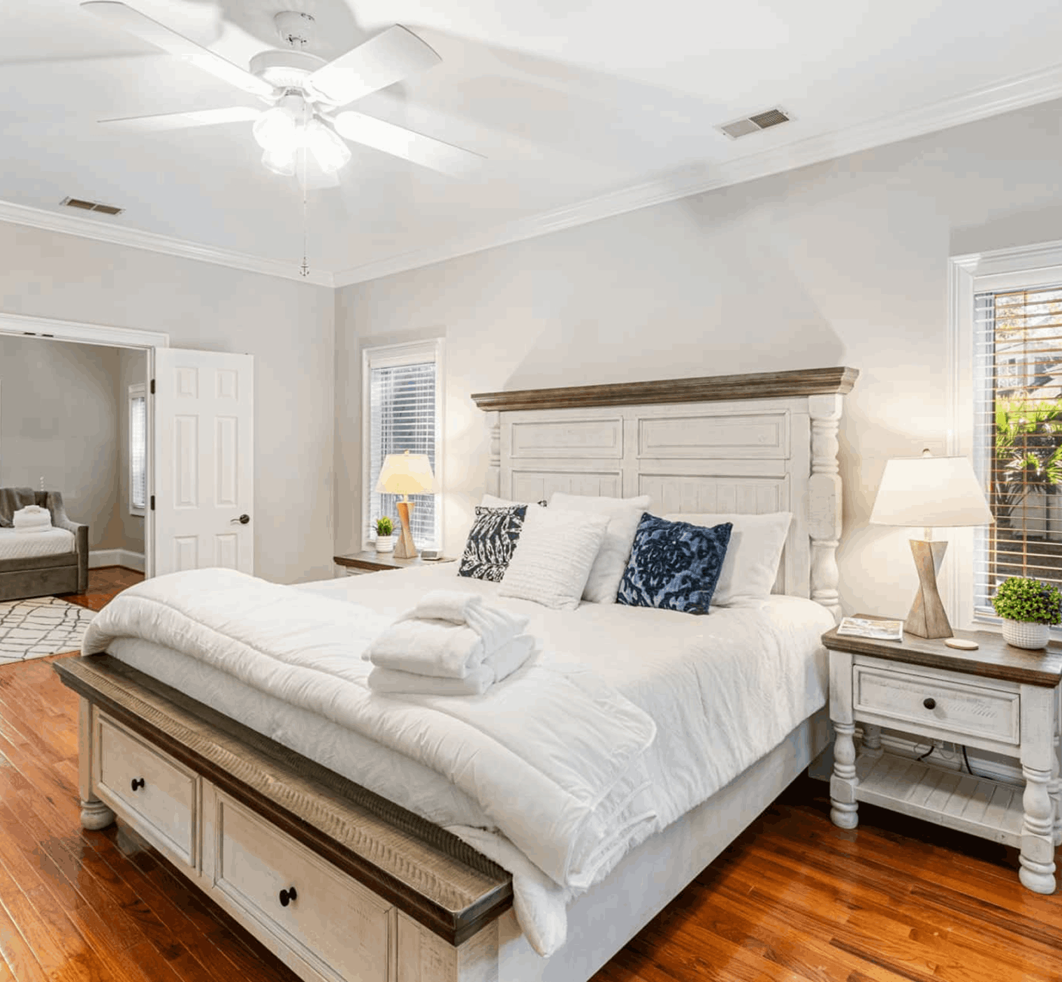 Bright bedroom with a large bed, white bedding, decorative pillows, two nightstands with lamps, a ceiling fan, and wooden flooring. A doorway leads to an adjoining room.