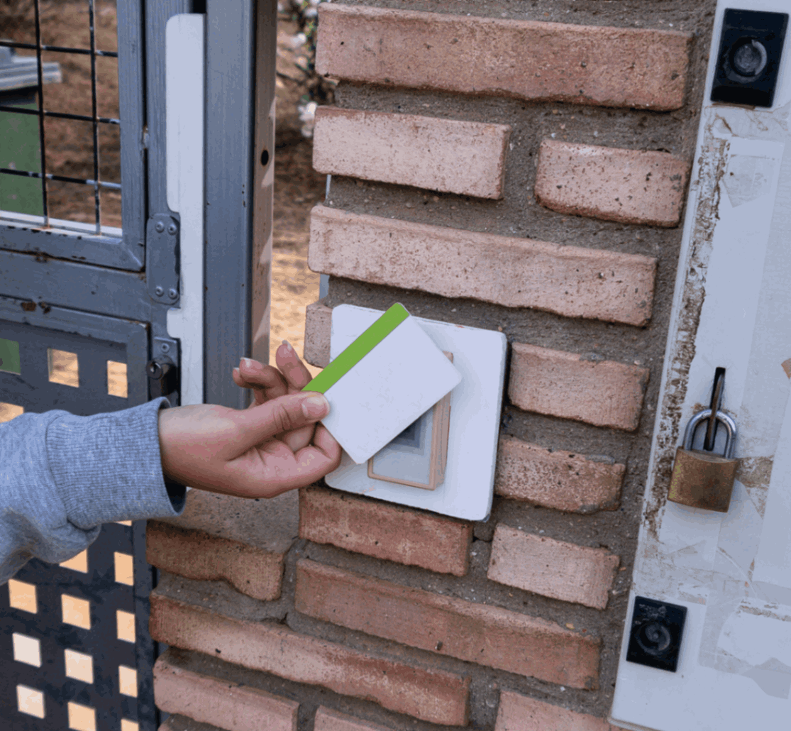 A person holds an access card to a card reader mounted on a brick wall next to a metal gate.